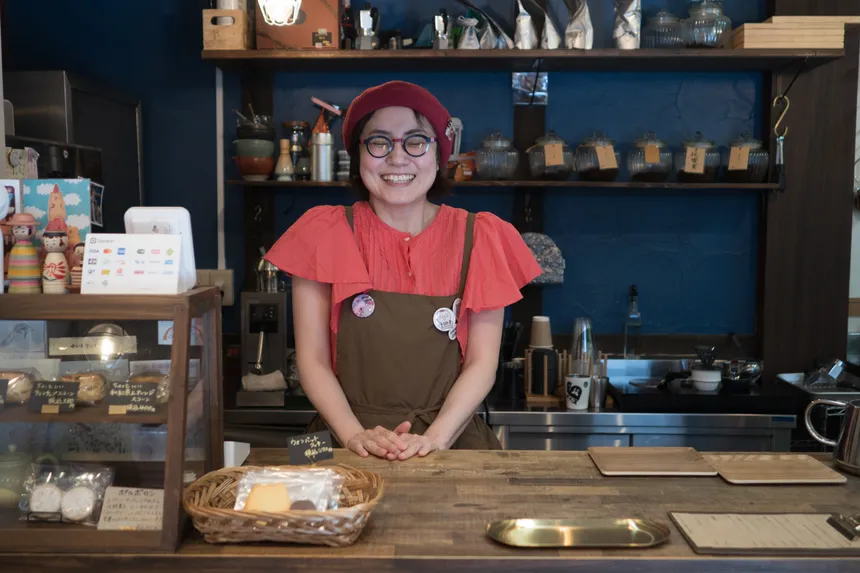 Rie-san sonríe detrás del mostrador de Suzumesya, con una blusa y un gorro rojos, ante una pared azul y estantes con tarros.