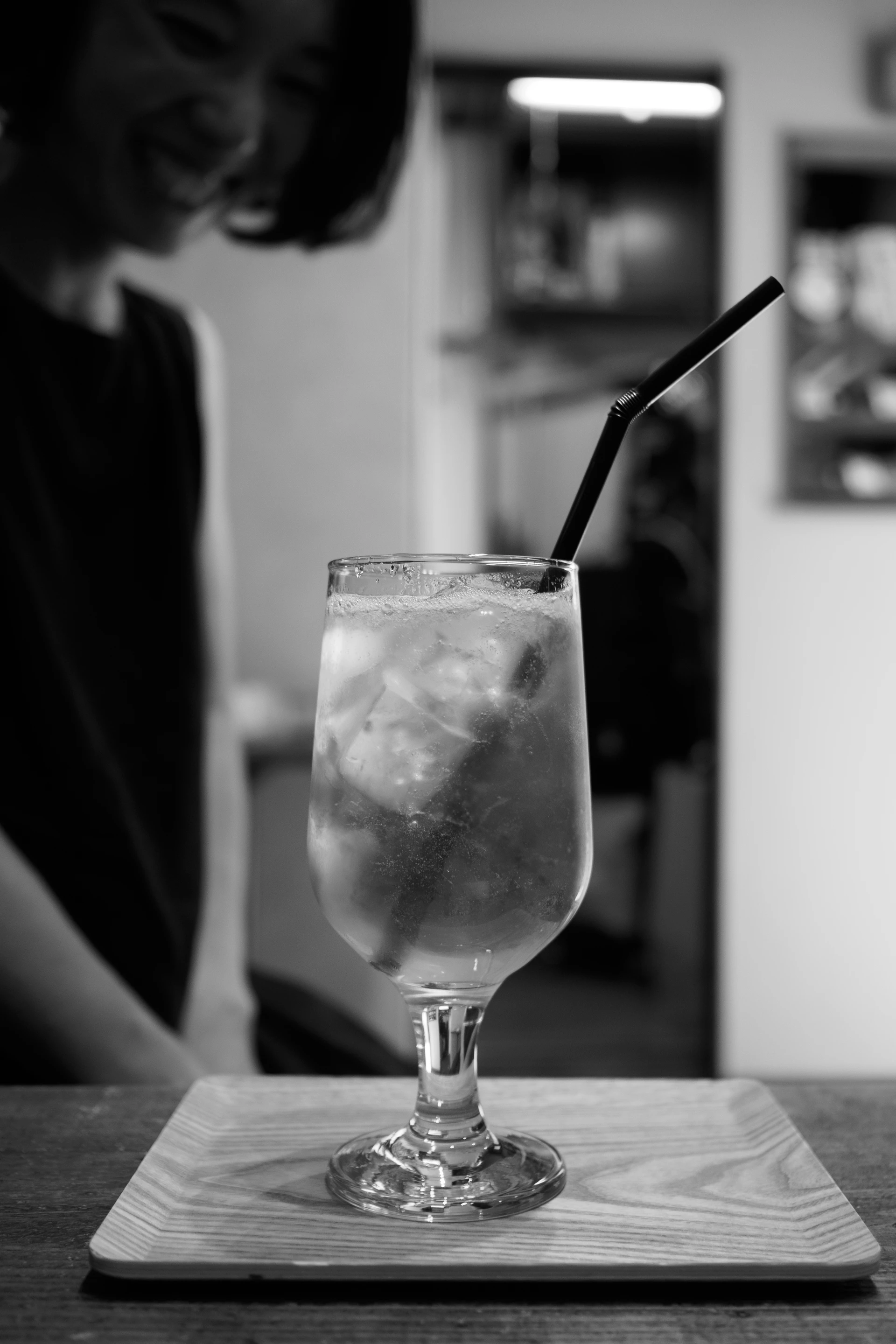 Una persona sonríe desenfocada al fondo, junto a un vaso helado con pajita en primer plano.