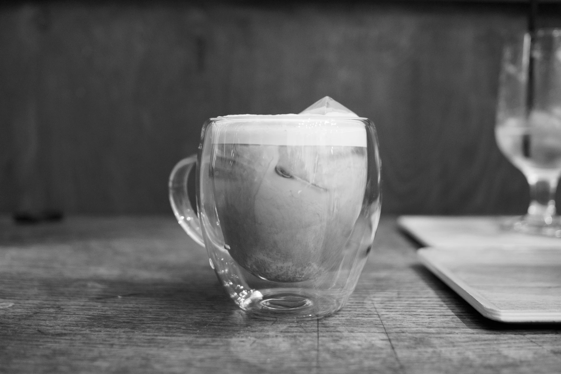 Una taza de latte con la espuma en pico sobre una mesa de madera, con un vaso al fondo.